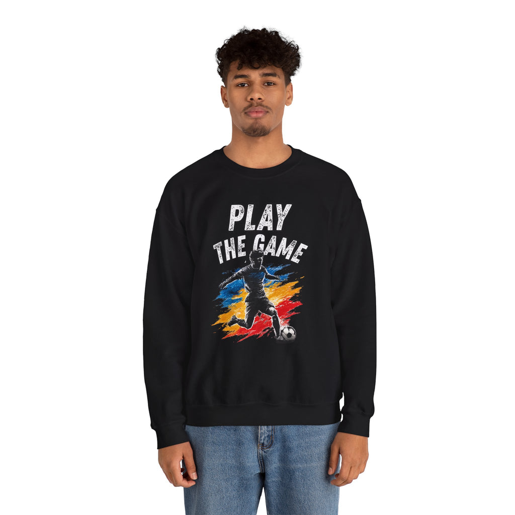 A man wearing a black crewneck sweatshirt with a graphic print that reads 'PLAY THE GAME' above a soccer player silhouette over a red, yellow, and blue paint-splatter design. He is wearing blue jeans against a plain white background.