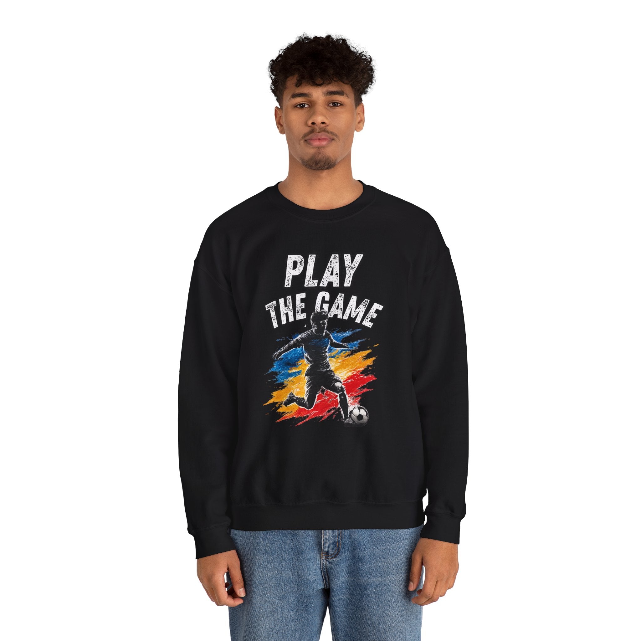 A man wearing a black crewneck sweatshirt with a graphic print that reads 'PLAY THE GAME' above a soccer player silhouette over a red, yellow, and blue paint-splatter design. He is wearing blue jeans against a plain white background.
