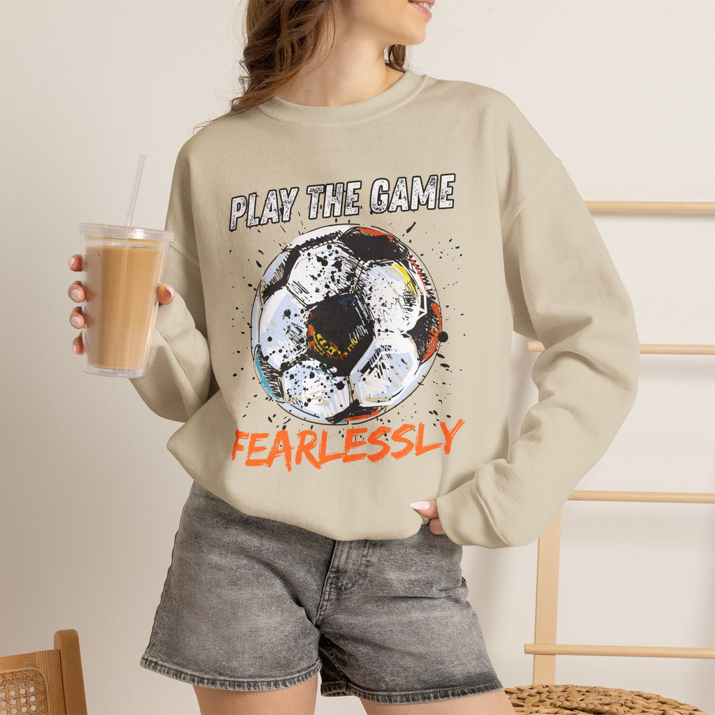 A woman wearing beige sweatshirt with dirty soccer ball graphic and play the game fearlessly text on a white background