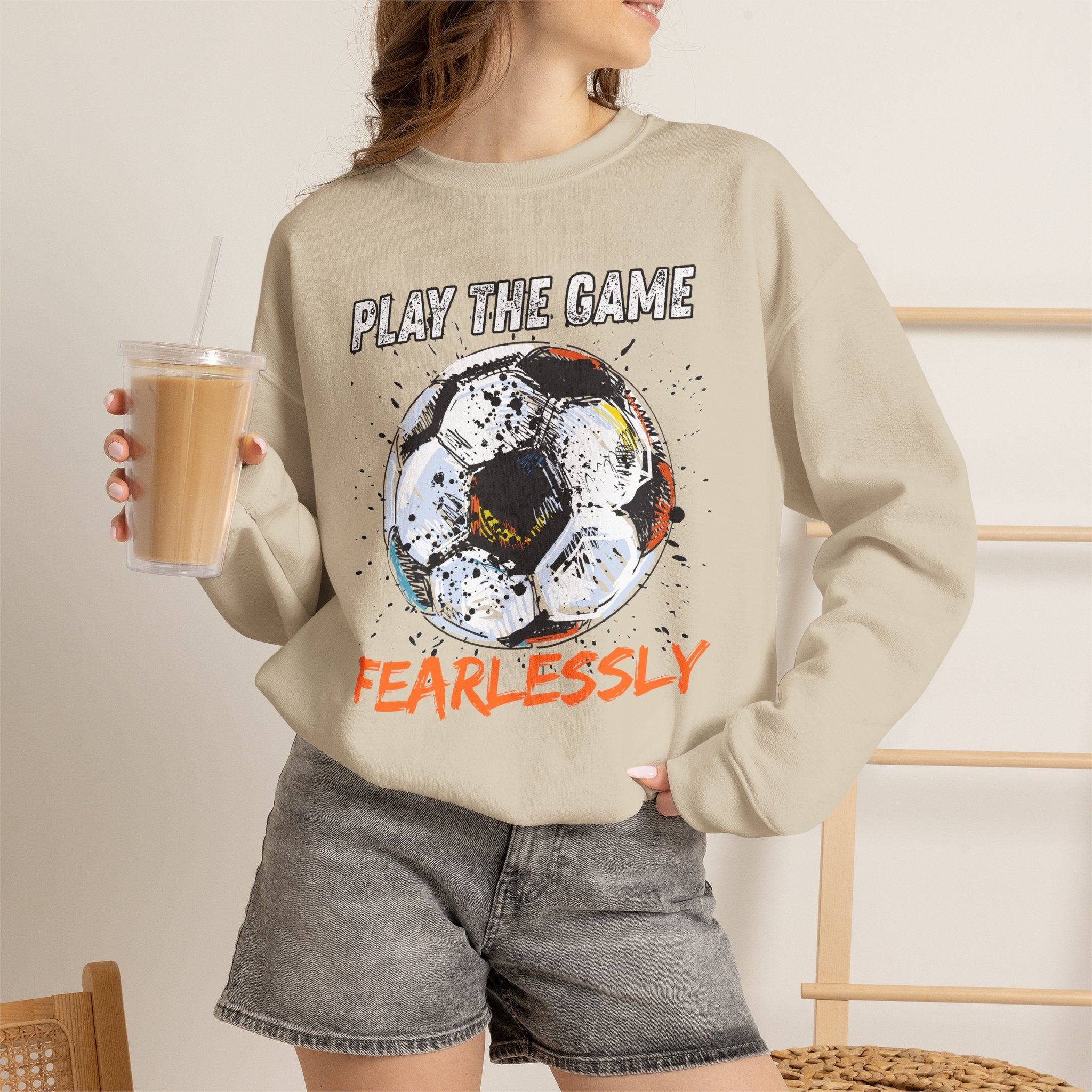 A woman wearing beige sweatshirt with dirty soccer ball graphic and play the game fearlessly text on a white background