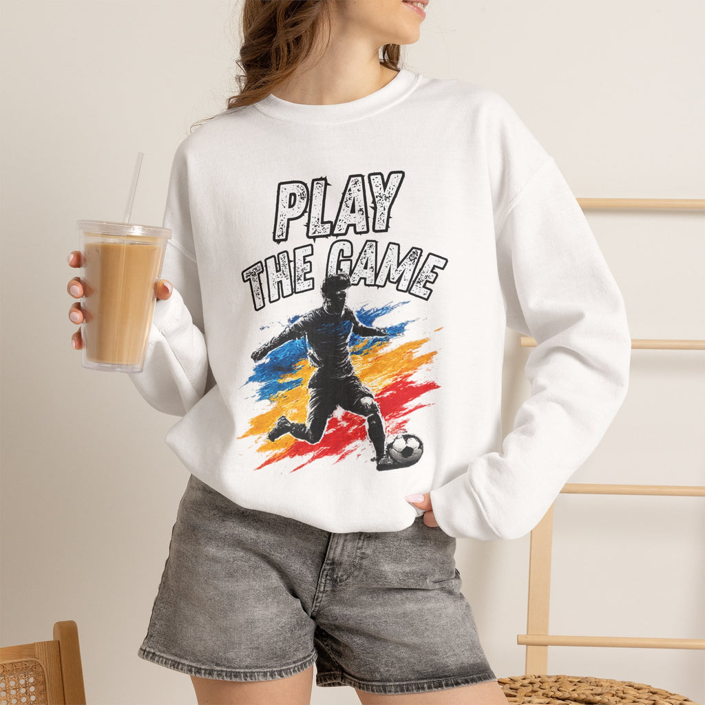 A woman wearing a white crewneck sweatshirt with a graphic print that reads 'PLAY THE GAME' above a soccer player illustration, paired with gray denim shorts and holding an iced coffee.