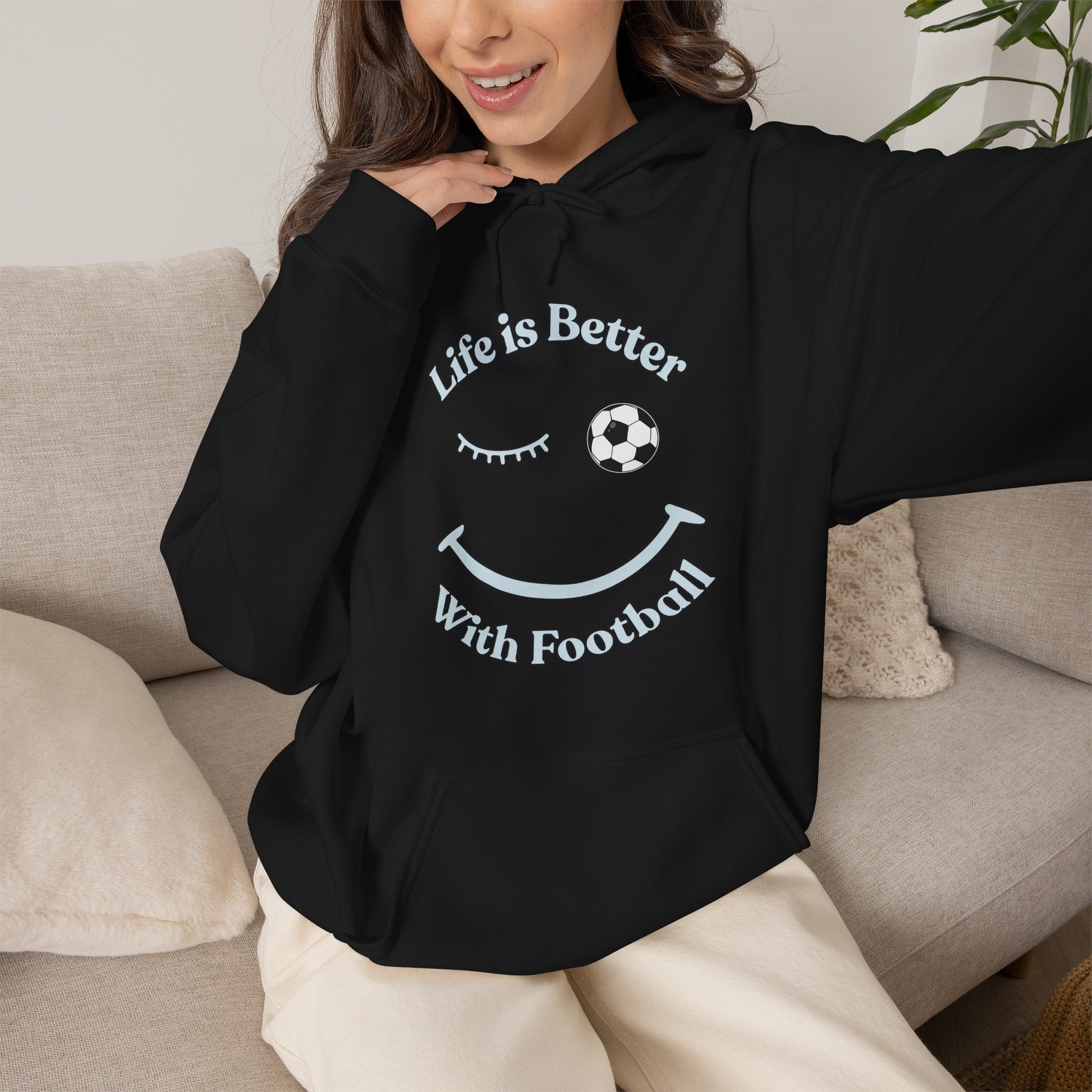 Person wearing a black hoodie with 'Life is Better With Football' text and smiley face design, sitting on a couch.