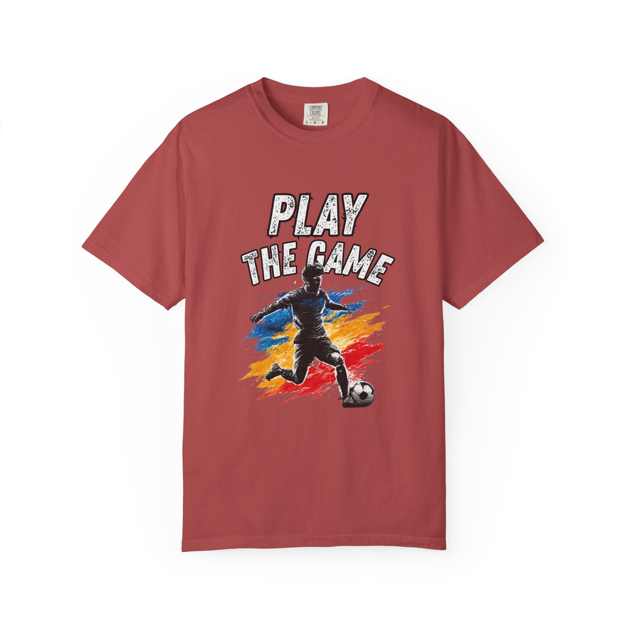 A short-sleeve crewneck t-shirt in a dusty rose or muted brick red color, featuring a graphic print on the front with the text 'PLAY THE GAME' above an illustration of a soccer player kicking a ball with colorful paint strokes.