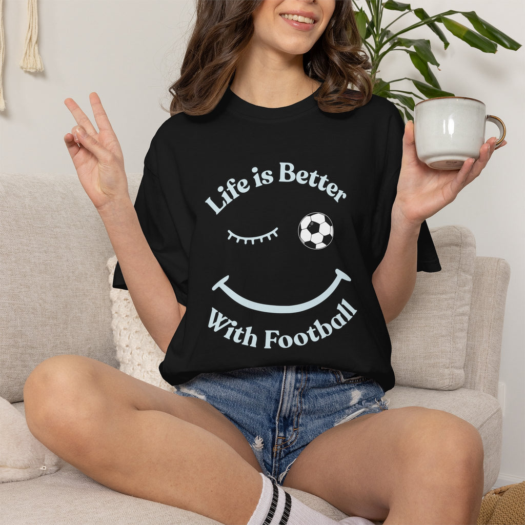 Person wearing a black t-shirt with 'Life is Better With Football' text and smiley face design, holding a mug.