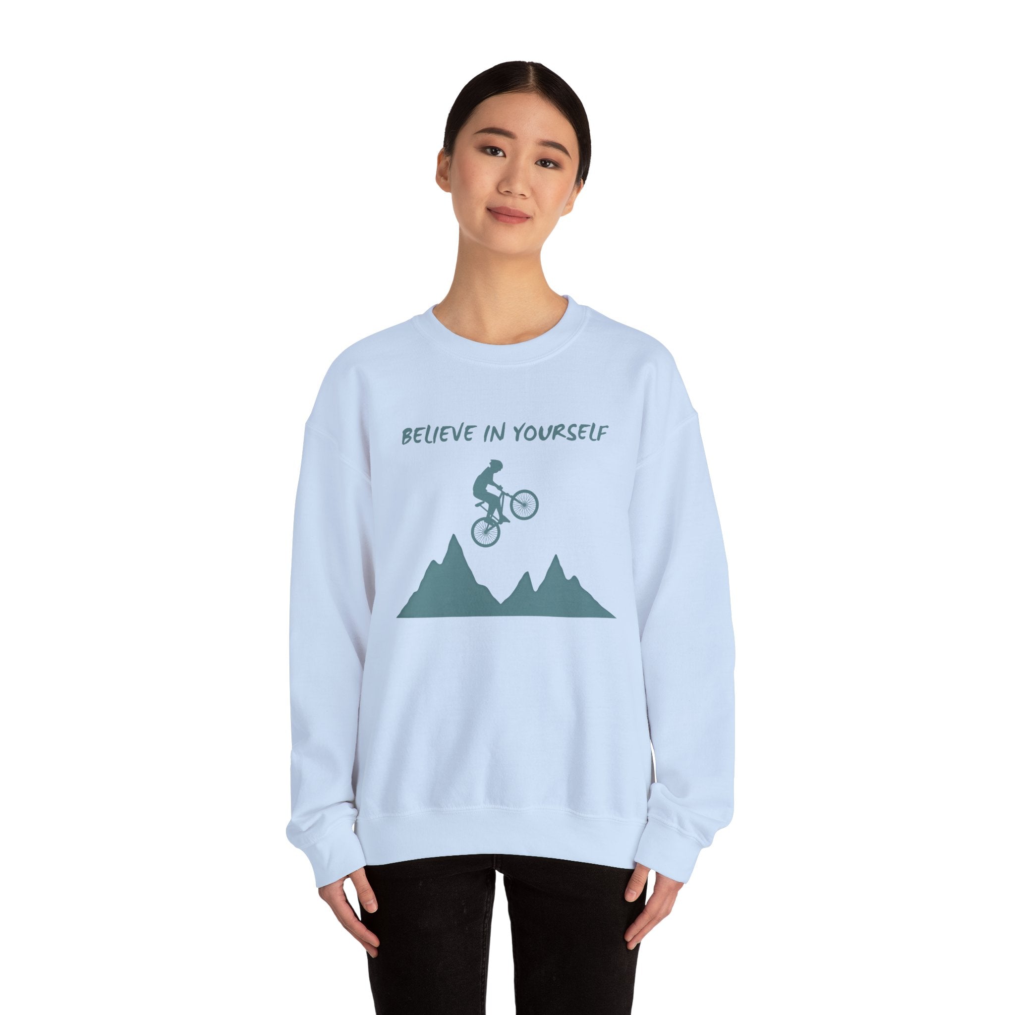 Light blue sweatshirt with mountain and bicycle graphic and 'Believe in Yourself' text worn by a person on a white background