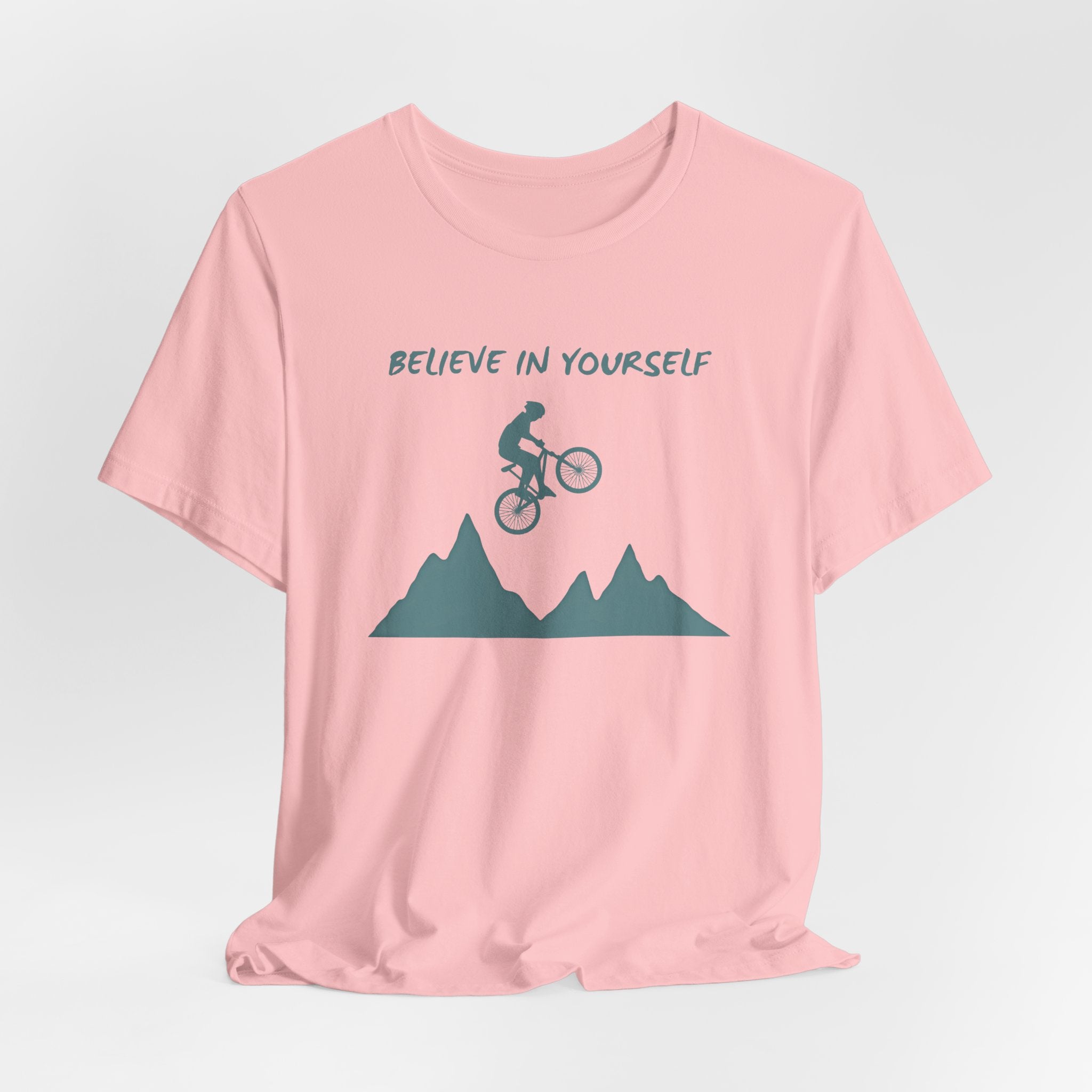 Pink t-shirt with mountain bike graphic and 'Believe in Yourself' text on a light gray background