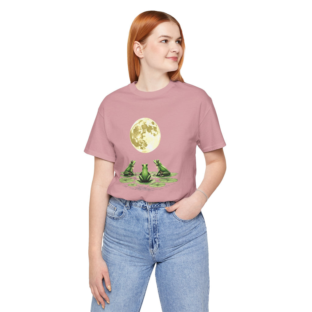 Person wearing a pink t-shirt with a graphic of three frogs and a moon, standing against a white background.